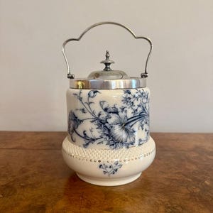 Puede incluir: Un tarro de cerámica blanco con un estampado floral azul, tapa y asa plateadas. El tarro tiene una base decorativa y una forma redondeada. El asa es arqueada. El diseño floral azul cubre el cuerpo del tarro.