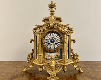 May include: A gold-toned, ornate clock with a white porcelain face and Roman numeral hour markers. The clock has a decorative design with floral motifs and a cherub figure on the face. The clock is sitting on a wooden surface.