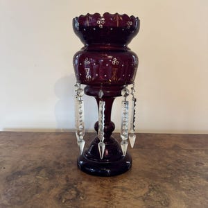 Könnte beinhalten: Eine dunkelrote Glasvase mit gewelltem Rand und weißen und goldenen Verzierungen. Die Vase hat hängende Kristallprismen und steht auf einem runden Sockel. Die Vase steht auf einer Holzoberfläche.