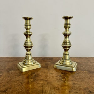May include: A pair of antique brass candlesticks with a fluted design and square bases. The candlesticks are on a wooden surface.