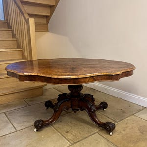 May include: An antique, oval wooden table with a dark brown finish. The table has a decorative base with carved details and claw feet. The tabletop is approximately 100 cm in diameter. The table is placed on a tiled floor.