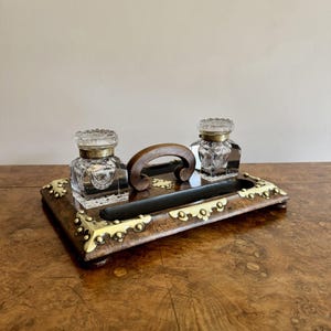 Peut inclure: Un ensemble de bureau ancien en bois avec deux encriers en verre, chacun avec un couvercle décoratif. L'ensemble comprend un porte-plume et une poignée en bois. Le plateau est orné de détails en laiton.