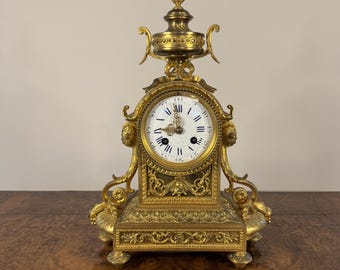 May include: A gold-toned mantle clock with ornate details and a white face. The clock has a decorative top and sits on a wooden surface.