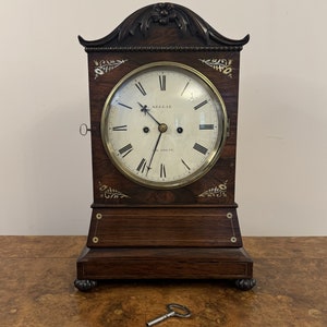 Puede incluir: Un reloj de chimenea antiguo de madera con una esfera blanca y agujas negras. El reloj tiene un borde de latón y una cerradura en el lateral. El reloj está sobre una superficie de madera con una llave delante.