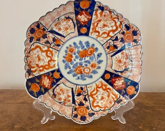 May include: Decorative ceramic plate with a scalloped edge. The plate features a central floral design in orange and blue, surrounded by intricate patterns and motifs in the same color scheme. The plate is displayed on a clear stand.