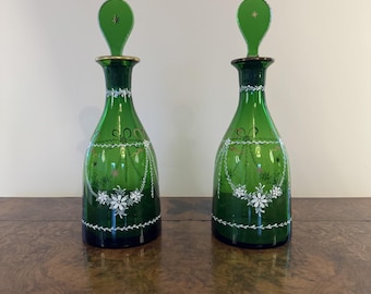 May include: A pair of green glass decanters with white floral designs and gold accents. The decanters have a round body and a long, thin neck. The stoppers are also green glass and have a star design.