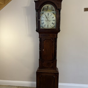 Puede incluir: Un reloj de pie alto de madera oscura con una esfera detallada y una escena pintada en la parte superior. El reloj tiene una base rectangular y una parte superior decorativa con un diseño curvo. La esfera del reloj tiene números romanos y manecillas ornamentadas.