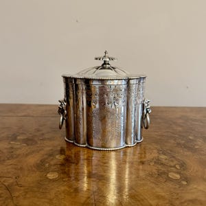 May include: A silver sugar bowl with a lid and two handles. The bowl is decorated with an intricate design and has a beaded edge.