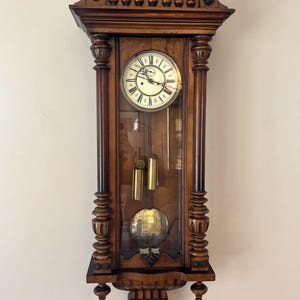 Könnte beinhalten: Eine antike Wanduhr aus Holz mit Glasfront. Das Zifferblatt ist weiß mit schwarzen römischen Ziffern und Zeigern. Die Uhr hat ein Pendel und Gewichte, die durch das Glas sichtbar sind. Die Uhr ist braun mit dekorativen Schnitzereien.