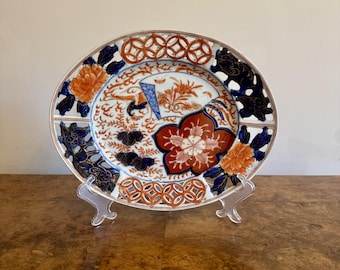 May include: An oval ceramic plate with a detailed floral design. The plate features orange, red, blue, and white colors. The rim has a geometric pattern. The plate is displayed on a clear plastic stand, showcasing its intricate artwork.