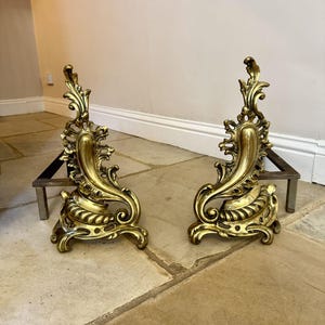 Pode incluir: Um par de tições de lareira em metal dourado com designs ornamentados de volutas curvas. Cada tição possui uma placa frontal decorativa alta com detalhes intrincados e uma barra de metal horizontal resistente que se estende para trás.