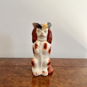 Puede incluir: Un jarro de cerámica con forma de perro marrón y blanco con un sombrero negro y dorado. El perro tiene orejas marrones, un cuerpo blanco y patas marrones. El jarro tiene un collar dorado alrededor del cuello.