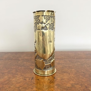 May include: A gold metal vase made from a repurposed artillery shell. The vase has a decorative design of leaves and a shield with an inscription.