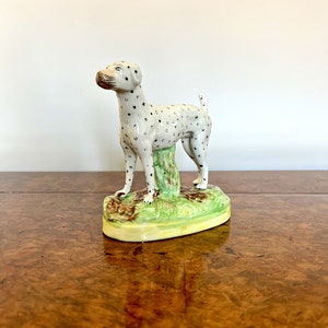 Op de afbeelding: Een wit keramiek beeldje van een Dalmatiër met zwarte vlekken, staand op een groene en bruine basis.