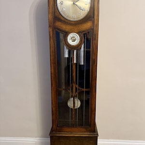 Könnte beinhalten: Eine antike Standuhr aus dunklem Holz. Das runde Zifferblatt zeigt römische Ziffern. Die Glastür gibt den Blick auf Pendel und Gewichte frei. Die Uhr steht auf einem rechteckigen Sockel.