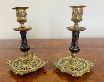 May include: A pair of ornate gold-toned brass candlesticks with blue and gold enamel detailing. The candlesticks have a square base with intricate scrollwork and a round top with a scalloped edge.