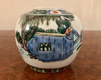 Puede incluir: Un jarrón de porcelana blanca con una escena pintada en azul, verde y amarillo de una persona de pie en un balcón con un árbol y un puente. El jarrón tiene una banda de rayas verdes, rojas y amarillas alrededor de la parte inferior.
