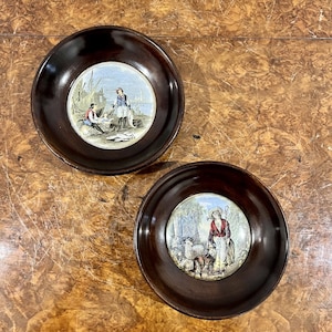 Puede incluir: Dos posavasos de madera marrón oscuro con un borde elevado y un inserto circular. El inserto presenta una escena pintada colorida. Un posavasos muestra a un hombre pescando en un bote con otro hombre en el fondo. El otro posavasos muestra a una mujer con un bastón y ovejas en un entorno boscoso.