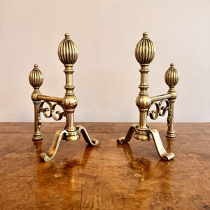 Puede incluir: Un par de morillos antiguos de latón. Cada morillo presenta un remate decorativo y acanalado sobre un soporte vertical, conectado por una barra horizontal con adornos en espiral. Los morillos tienen pies curvos.