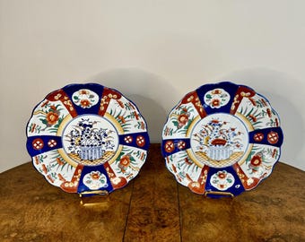 May include: Two decorative plates with scalloped edges, featuring a floral design in a blue, red, and gold color scheme. Each plate has a central floral arrangement in a basket. The plates are displayed on a wooden surface.