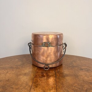 May include: A copper bucket with a hinged lid and a metal handle. The bucket is sitting on a wooden table.