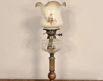 May include: A vintage brass oil lamp with a clear glass shade and a white glass chimney with a floral design. The lamp has a decorative base with ornate details.