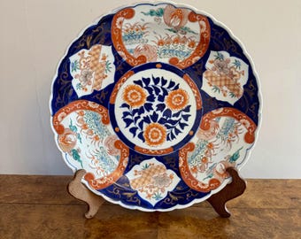 May include: A decorative ceramic plate with a scalloped edge. The plate features a dark blue border with orange and white floral and bird designs. The center has a floral pattern in orange and blue. The plate is displayed on a wooden stand.
