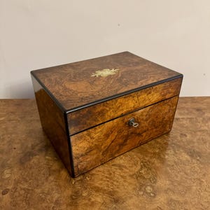 Puede incluir: Una caja de madera con acabado de madera de raíz y un pequeño ojo de cerradura de latón. La parte superior de la caja tiene una incrustación decorativa de un diseño floral.