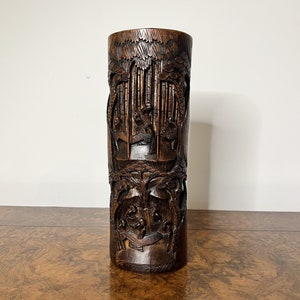 Può includere: Un vaso cilindrico alto, scolpito in bambù marrone scuro. Il vaso presenta sculture intricate di figure e alberi, creando una superficie dettagliata e testurizzata.