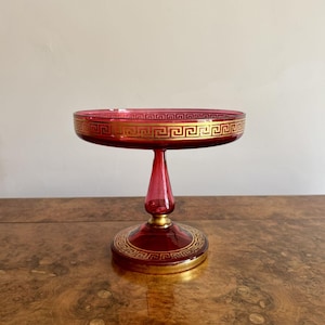 Puede incluir: Un plato con pedestal de vidrio rojo rubí con un diseño de greca dorada alrededor del borde y la base. El plato tiene un cuenco redondo y poco profundo y un tallo delgado, perfecto para servir postres o exhibir objetos pequeños.