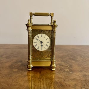 May include: A gold-toned brass mantel clock with a white face and black Roman numeral hour markers. The clock has a decorative engraved design and a handle on the top.