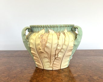 May include: A decorative vase with a light green rim and handles, featuring a design of cream-colored, feather-like shapes. The vase has a textured surface and is set on a wooden table.