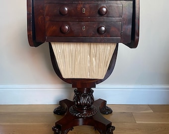 May include: A mahogany drop-leaf table with two drawers and a fabric-covered base. The table has a traditional design with turned legs and claw feet.