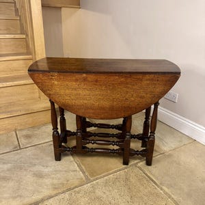 Puede incluir: Mesa antigua abatible de madera marrón oscuro. La mesa tiene una tapa semicircular y patas torneadas con una base de camilla detallada. La veta de la madera es visible. La mesa está sobre un suelo de piedra. Altura aproximada de 76 cm.