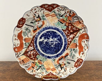 May include: A large, round, white ceramic plate with a scalloped edge. The plate is decorated with a colorful Japanese-style design featuring figures, trees, and geometric patterns in shades of blue, red, green, and gold.