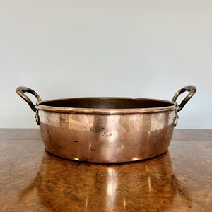 Puede incluir: Una olla redonda de cobre con dos asas de latón. La olla tiene una superficie brillante, ligeramente deslustrada.
