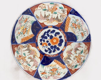 May include: A large, round, white porcelain plate with a blue and red floral design. The plate is decorated with a central floral motif and four smaller floral motifs around the edge. The plate is also decorated with gold accents.