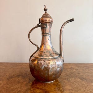 May include: Antique copper pitcher with a long spout and a hinged lid. The pitcher has a bulbous body and a curved handle. The metal has a weathered appearance with areas of discoloration, suggesting age and use. The pitcher is displayed on a wooden surface.