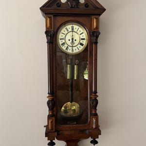 Puede incluir: Reloj de pared antiguo de madera con esfera clara, números romanos y tallas decorativas. El reloj tiene un péndulo y dos pesas. El reloj está montado en una pared de color claro.