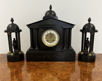 Könnte beinhalten: Eine schwarze Marmorkaminuhr mit zwei passenden schwarzen Marmor-Kerzenständern. Die Uhr hat ein Zifferblatt mit römischen Ziffern und goldenen Akzenten. Die Kerzenständer haben eine goldene Basis und eine schwarze Marmorsäule mit einer goldenen Spitze.