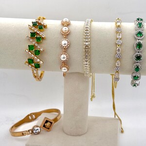 Puede incluir: Un conjunto de siete pulseras de oro y plata con piedras preciosas verdes y blancas. Las pulseras se muestran en un soporte blanco.