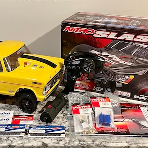 May include: A yellow and black toy truck with a black spoiler and a black stripe on the hood. The truck is next to a box that says "Nitro Slash" and various parts and accessories.