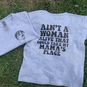 K&ouml;nnte beinhalten: Graues T-Shirt mit einer schwarz-wei&szlig;en Illustration eines Personenkopfes auf der Vorderseite und dem Text "Ain't a woman alive that could take my mama's place" auf der R&uuml;ckseite.