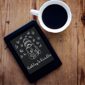 Puede incluir: Una lectora electrónica negra con una ilustración blanca de una persona leyendo un libro rodeada de mariposas. El texto "Ashley's Kindle" está escrito en la pantalla.