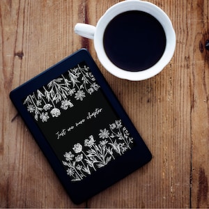 May include: A black e-reader with a white floral design and the text "Just one more chapter" on the screen. The e-reader is sitting on a wooden surface next to a white coffee cup.