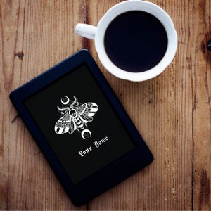 Puede incluir: Un lector electrónico negro con un gráfico blanco de una polilla con una calavera en la cabeza. La polilla está rodeada de un diseño geométrico con lunas crecientes. El texto "Your Name" está impreso en la pantalla.