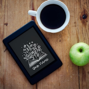 Könnte beinhalten: Ein schwarzer E-Reader mit einer weißen Umrandung eines Buches, aus dem Blumen wachsen. Der Text "Your Name" ist auf dem Bildschirm geschrieben. Eine Tasse Kaffee und ein grüner Apfel stehen auf einem Holztisch.