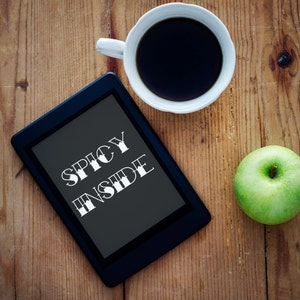 Op de afbeelding: Een zwarte e-reader met de tekst "Spicy Inside" op het scherm. De e-reader ligt op een houten tafel met een kop koffie en een groene appel.