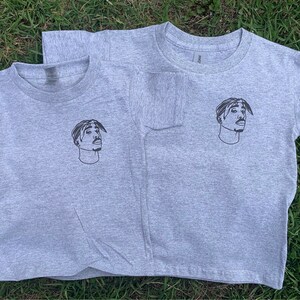 K&ouml;nnte beinhalten: Zwei graue T-Shirts mit einem Schwarzwei&szlig;bild des Rappers Tupac Shakur auf der Vorderseite.