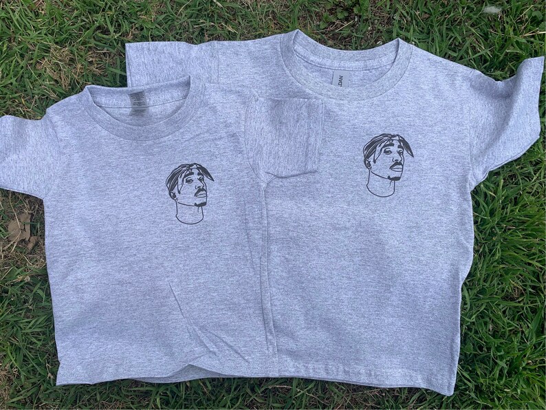 K&ouml;nnte beinhalten: Zwei graue T-Shirts mit einem Schwarzwei&szlig;bild des Rappers Tupac Shakur auf der Vorderseite.